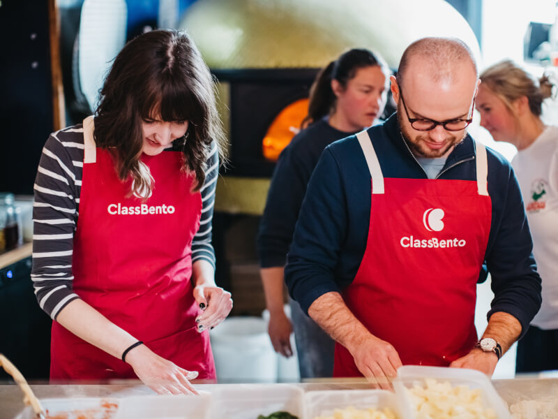 A couple wearing aprons making pizza at a cooking class.